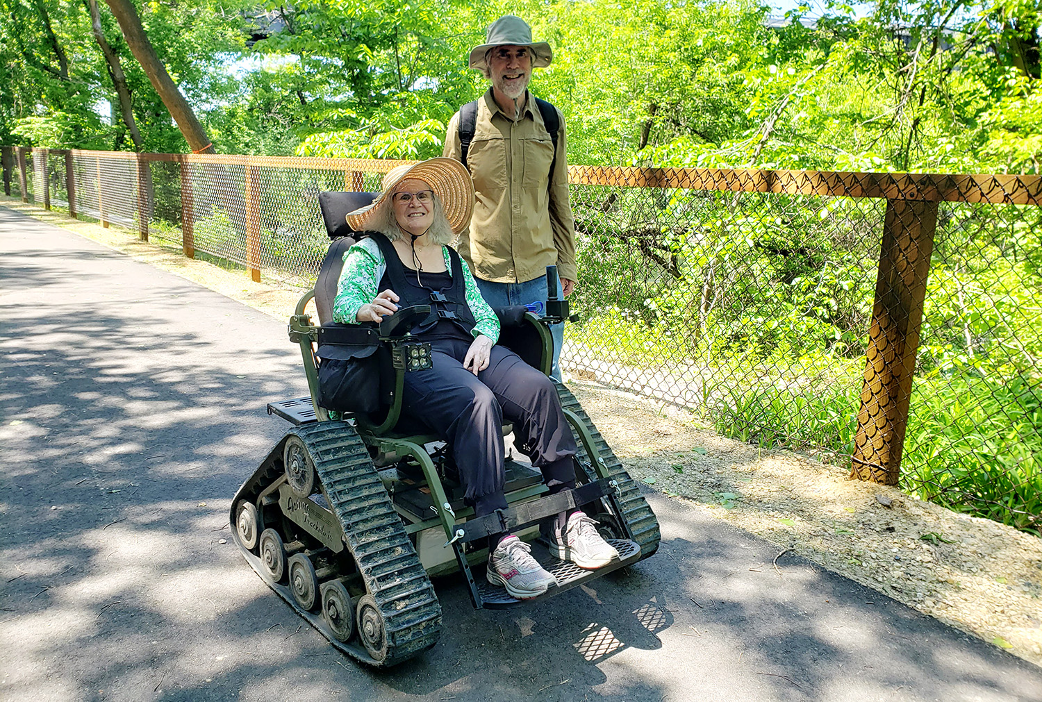 A woman in a motorized wheelchair designed with tank-like tracks is smiling, wearing a wide-brimmed straw hat, a green top, and black pants. Standing beside her is a man in a wide-brimmed hat, khaki shirt, and dark pants. They are outdoors on a sunny day near a fenced path with greenery in the background.